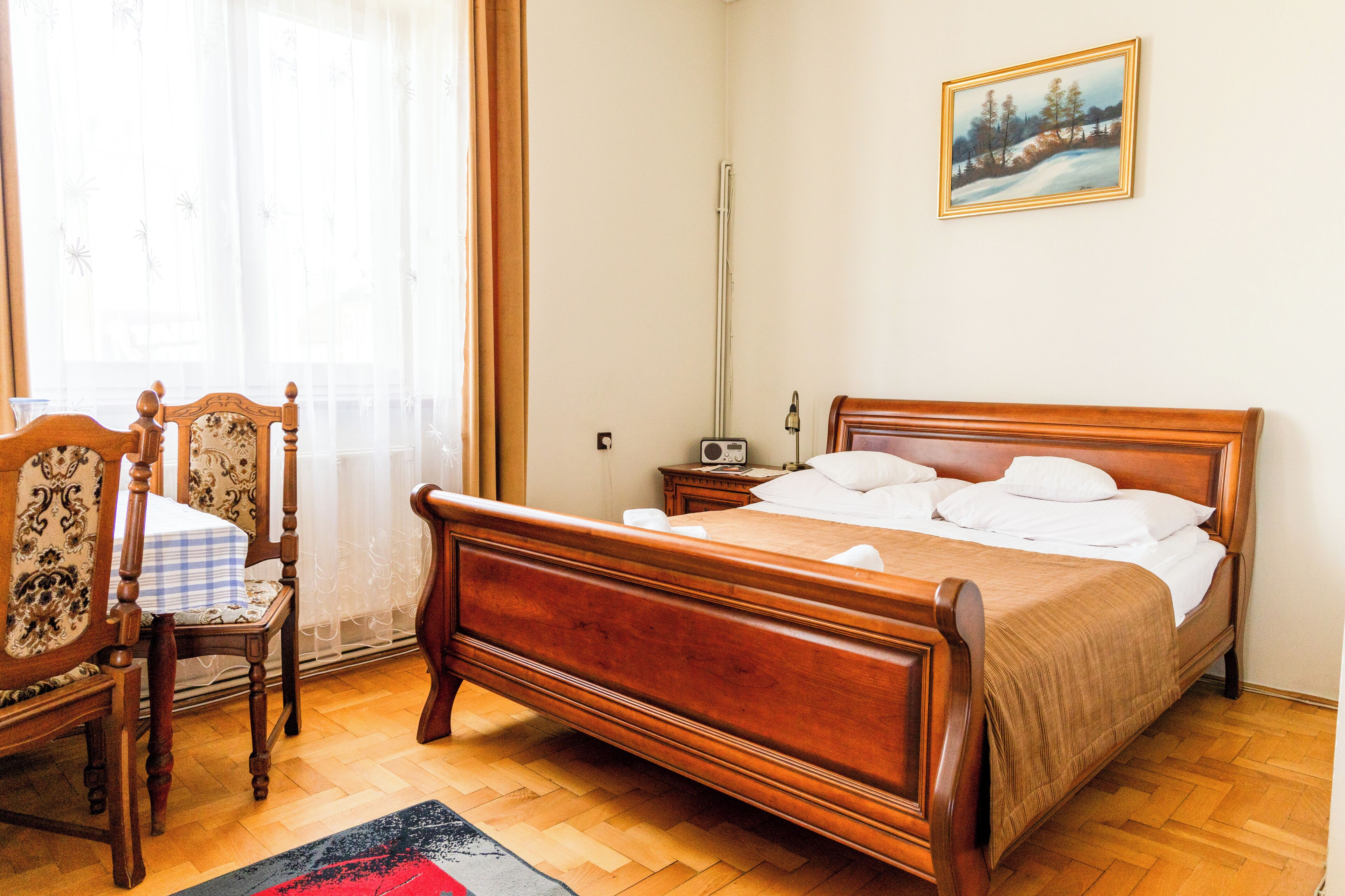 A bedroom with a wooden bed, a chair, a bedside table, a radio, and a painting on the wall.
