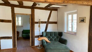 Une pièce avec un canapé vert recouvert de tissu, des poutres en bois, un lampadaire et, par une porte ouverte, on aperçoit une autre pièce où se trouve un autre canapé vert.