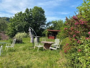 En rustik utomhusplats med sittmöbler, bestående av metallstolar och ett bord, omgiven av frodig grönska och blommande buskar.