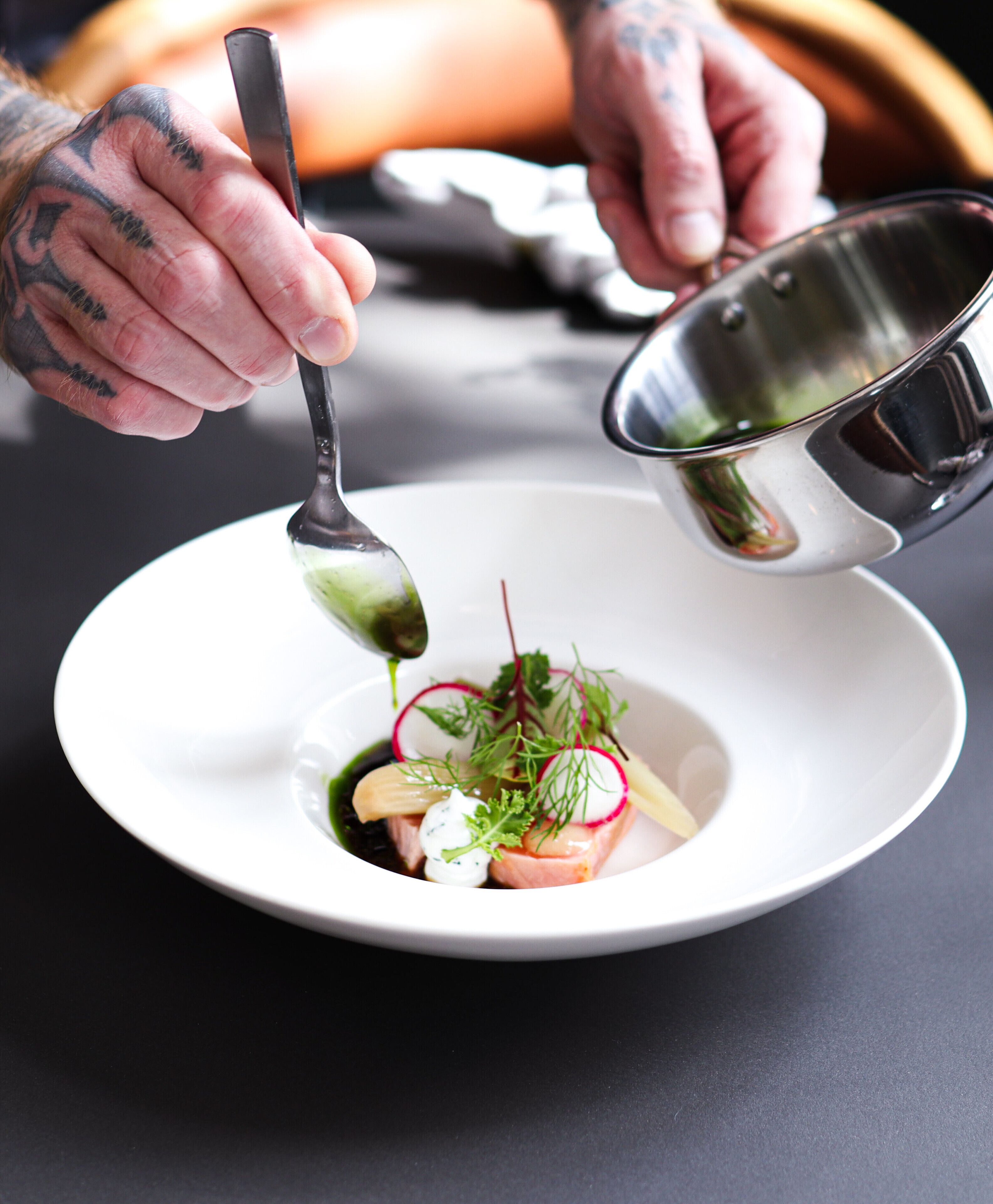 A person pouring a liquid from a metal bowl into a white plate with a spoon.