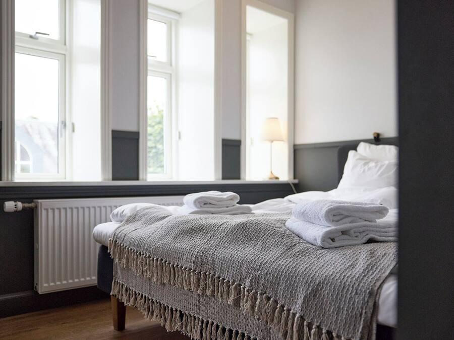Un letto rifatto con lenzuola bianche e una coperta grigia con frange, una lampada da comodino e vista su un edificio bianco attraverso una finestra.