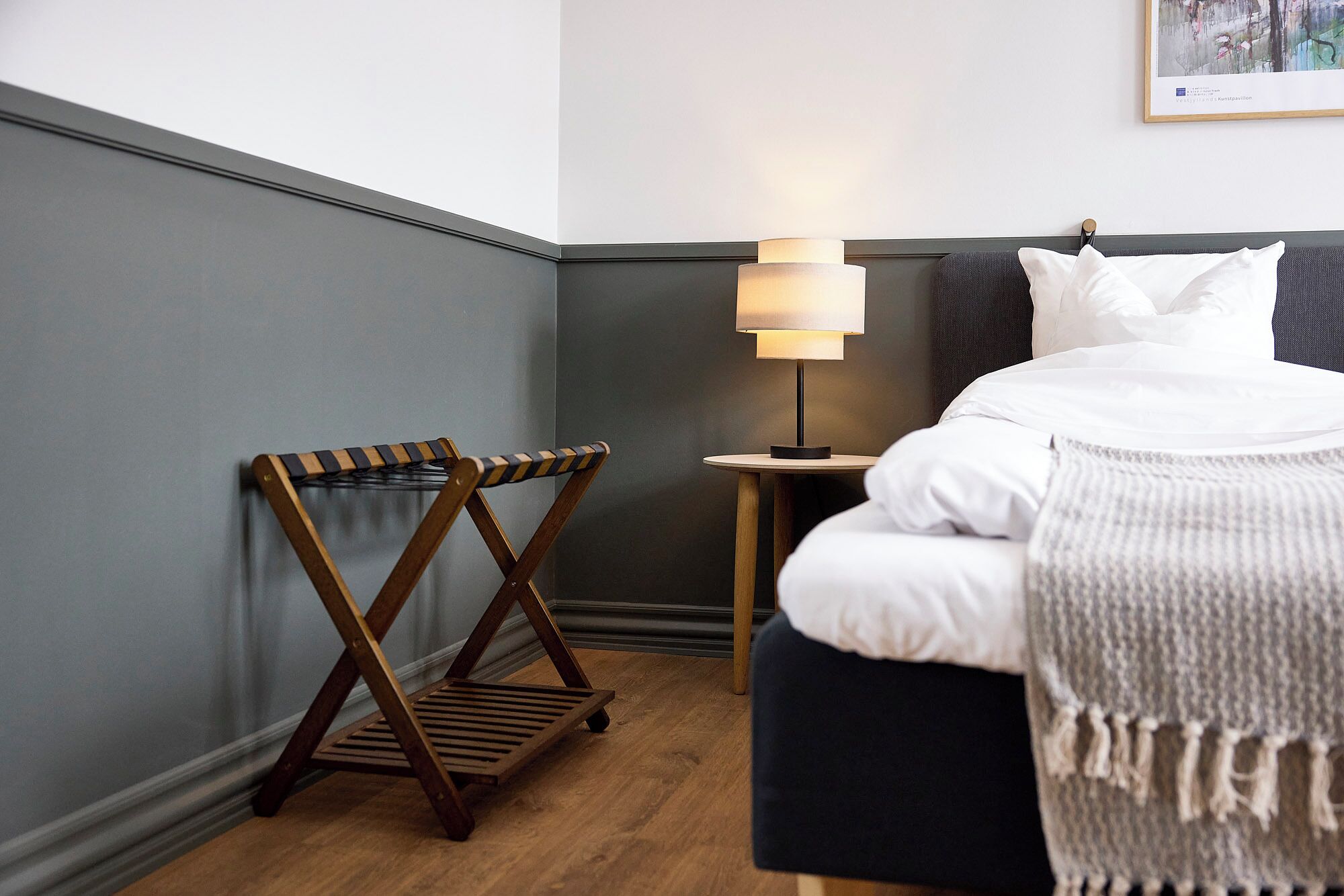 A hotel room with a bed, a bedside table with a lamp, a wooden chair, and a wall-mounted artwork.