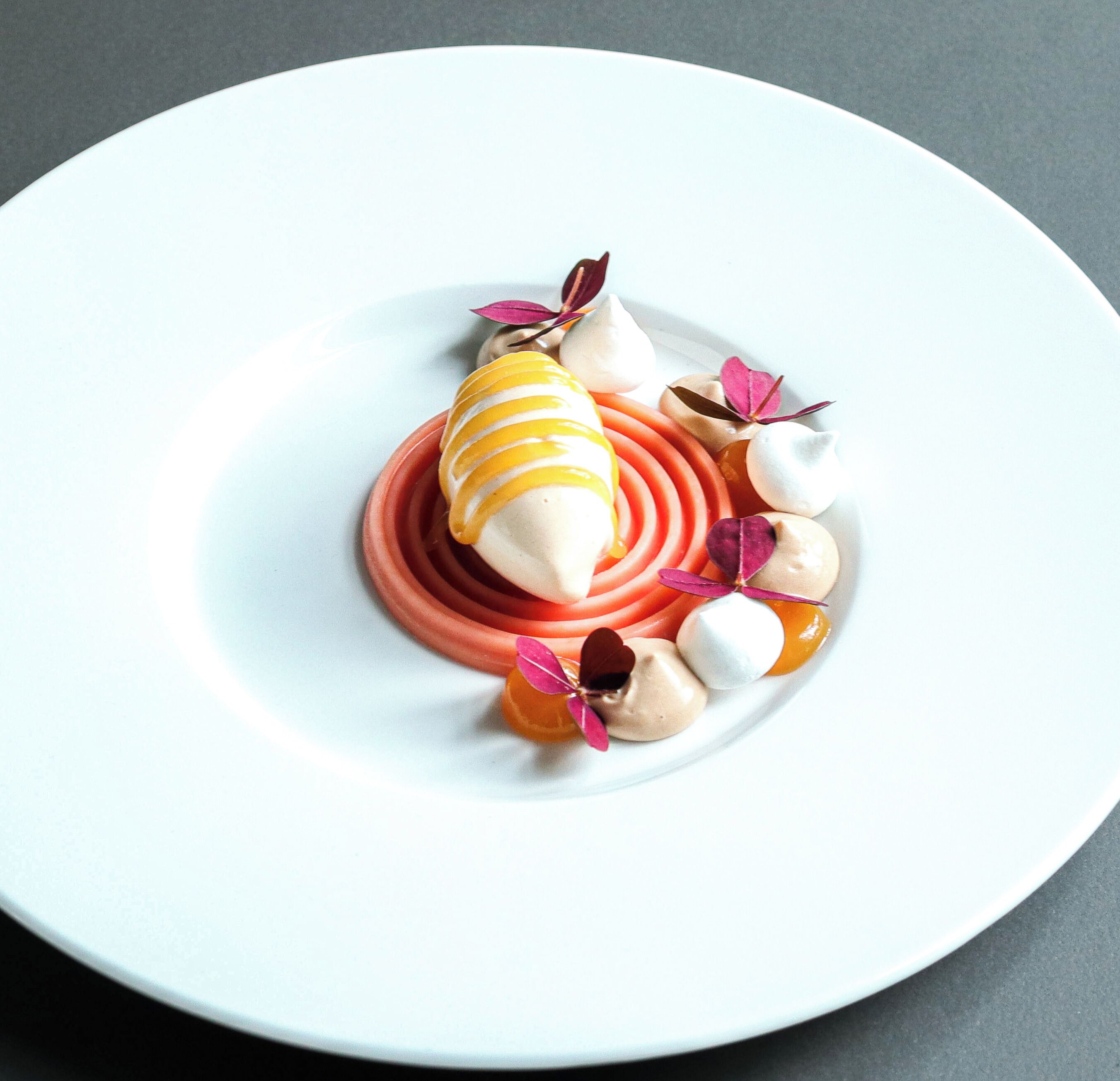Een wit bord met een dessert bestaande uit een ijsje met gele strepen, een roze saus in een spiraalvorm en kleine ronde meringues met roze blaadjes.