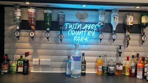 A bar area with a neon sign reading 'Twin Ays Country Park' and various liquor bottles. - Glan yr Afon Tri - Relax, Recuperate, Hot Tub (Welshpool)