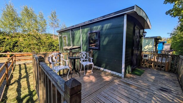 A wooden deck with two white metal chairs and a small table, a green building with a curved roof, and a wooden fence. - Glan yr Afon Tri - Relax, Recuperate, Hot Tub (Welshpool)