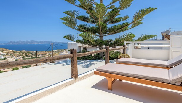 Un balcón con barandilla de madera, una silla de descanso blanca y vistas al mar y al paisaje distante.