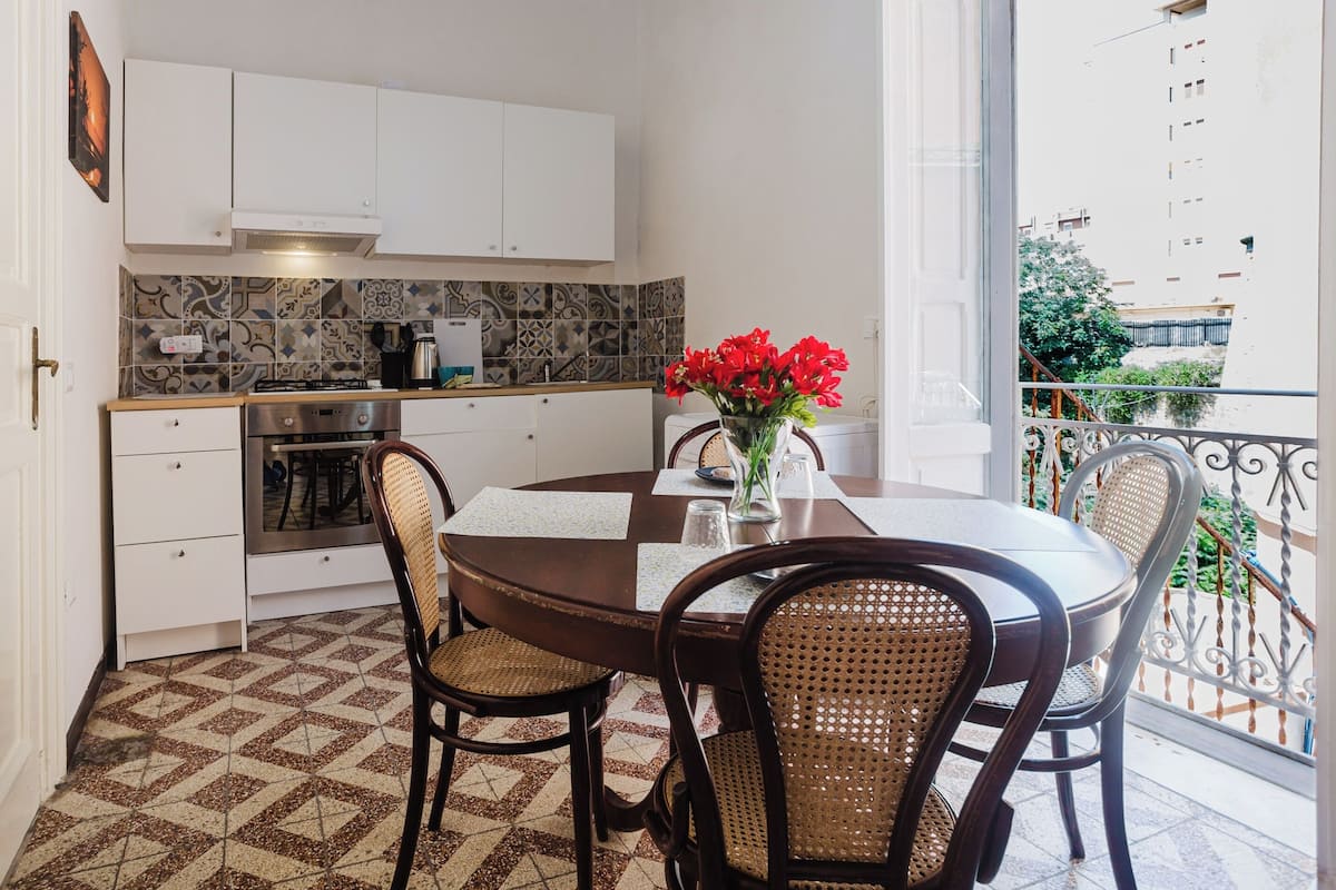 A compact kitchen with white cabinets, a built-in oven, and a tiled backsplash. A dining table with four chairs is set for a meal.