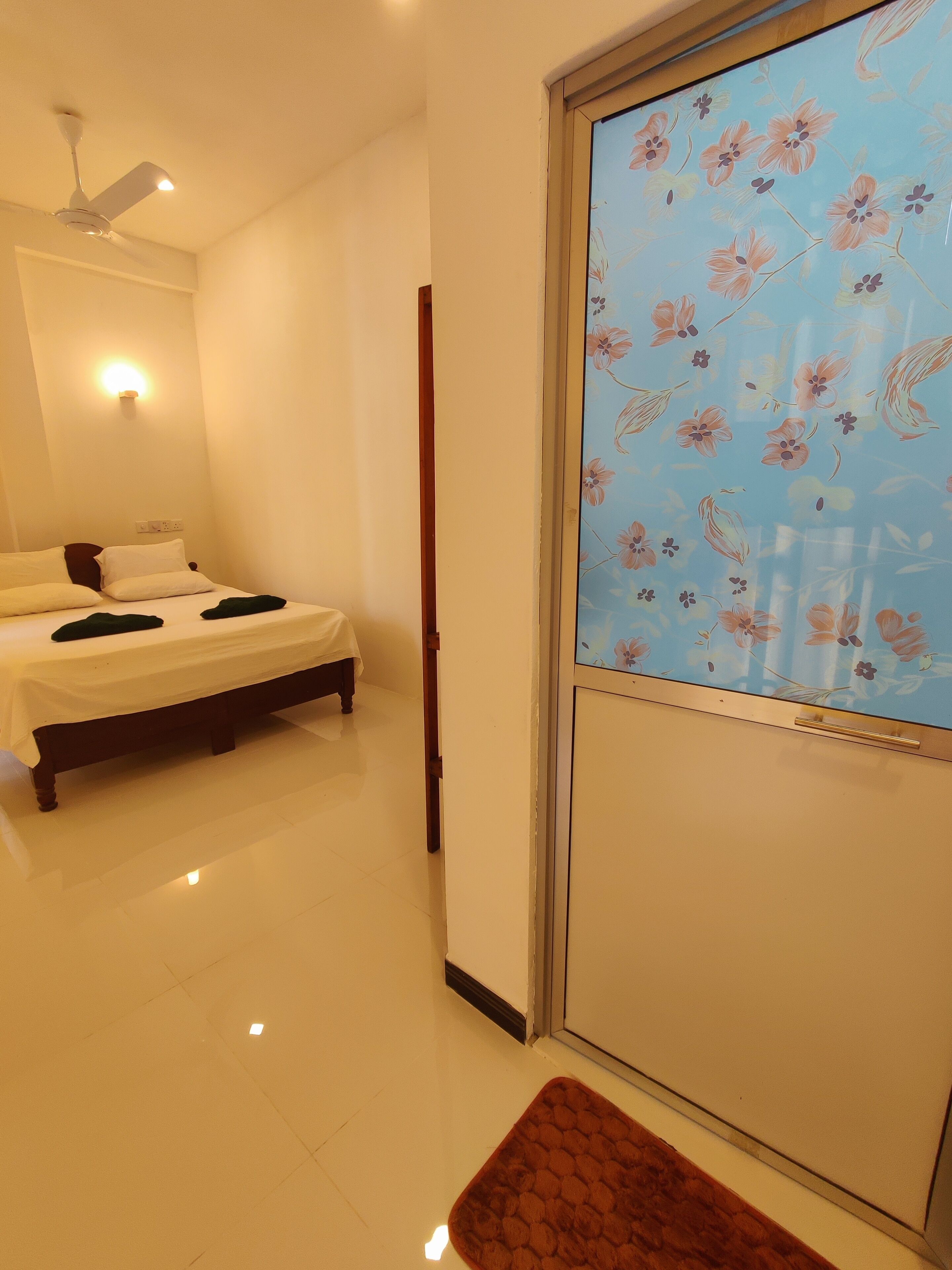 A hotel room with a bed, a ceiling fan, a wall-mounted light, and a door with a floral-patterned glass panel.