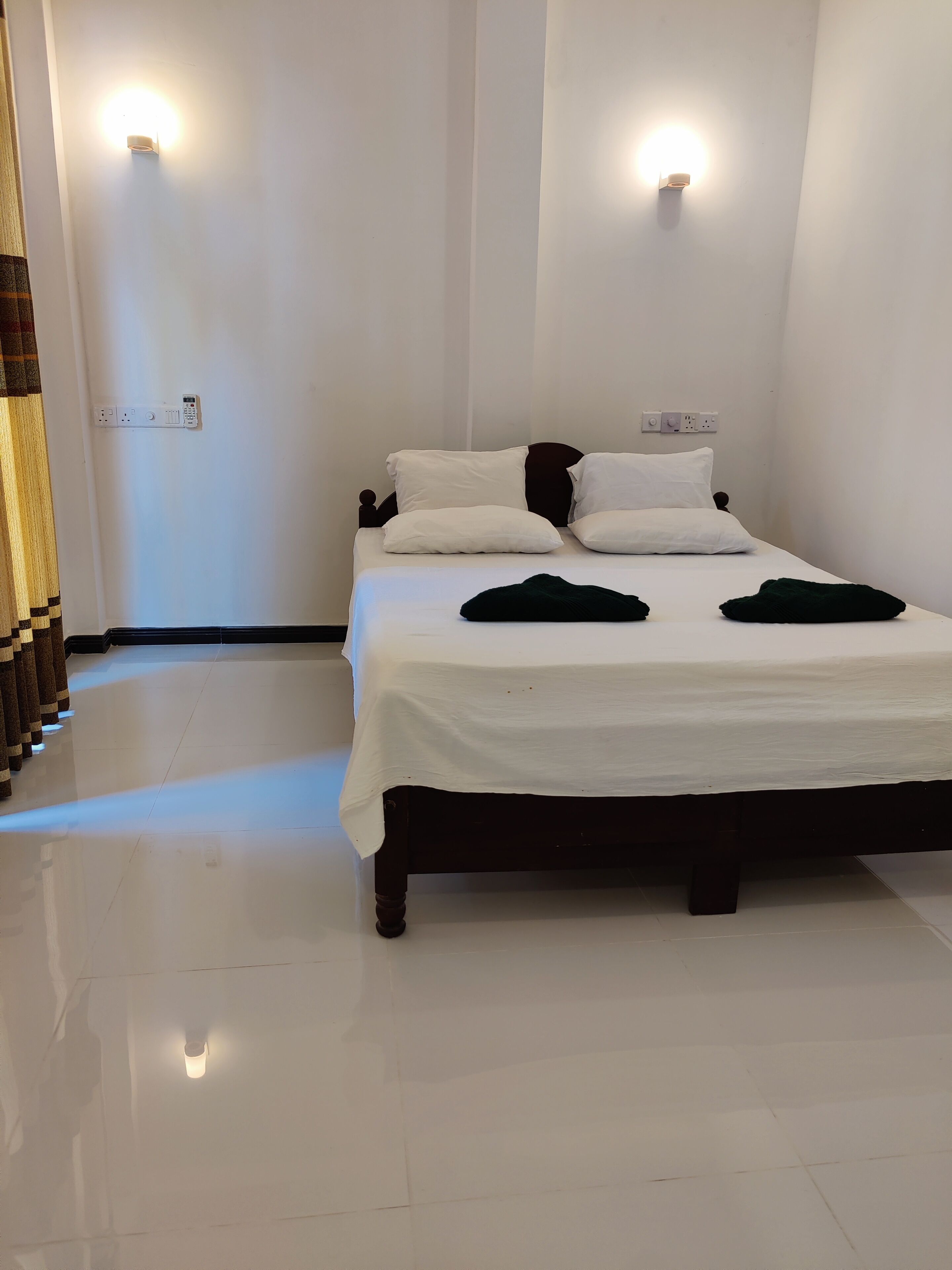 A neatly made bed with white linens and two dark throw pillows, a wall-mounted light fixture, and a power outlet on the wall.