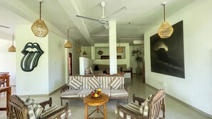 A spacious living area with a sofa, two wooden chairs, a small round table with a flower vase, a ceiling fan, and a large abstract painting on the wall. - Randi Guest House (Mirissa)