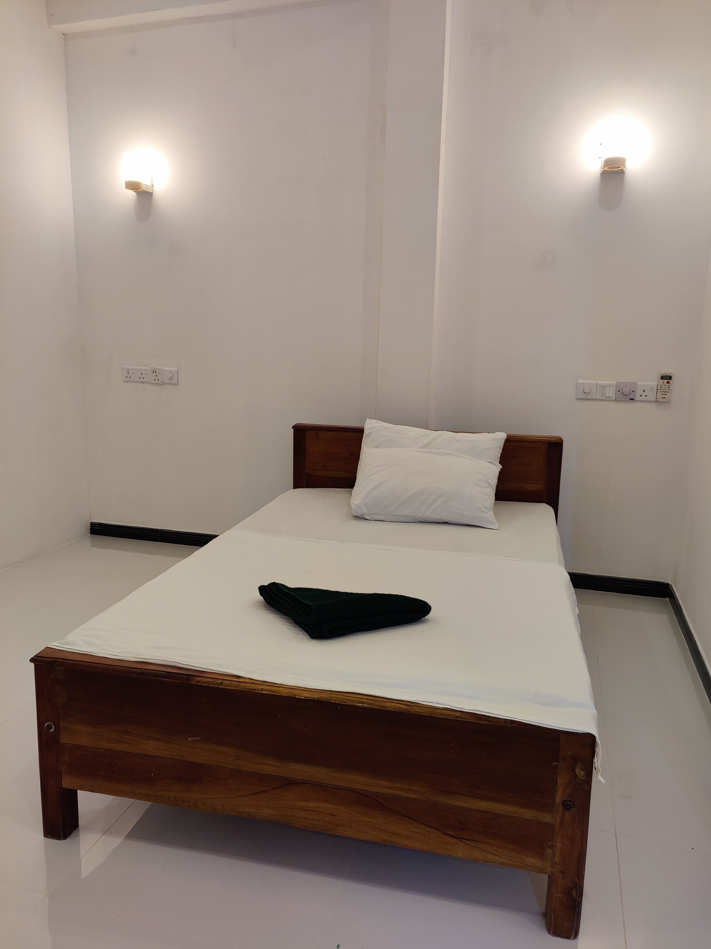 A single bed with a wooden frame and white bedding, two wall-mounted light fixtures, and a power outlet on the wall.