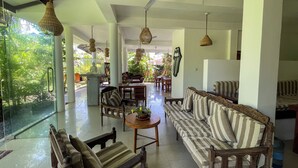A spacious indoor area with a large sofa, wooden chairs, and hanging lights. There is a glass wall with a view of greenery outside. - Randi Guest House (Mirissa)