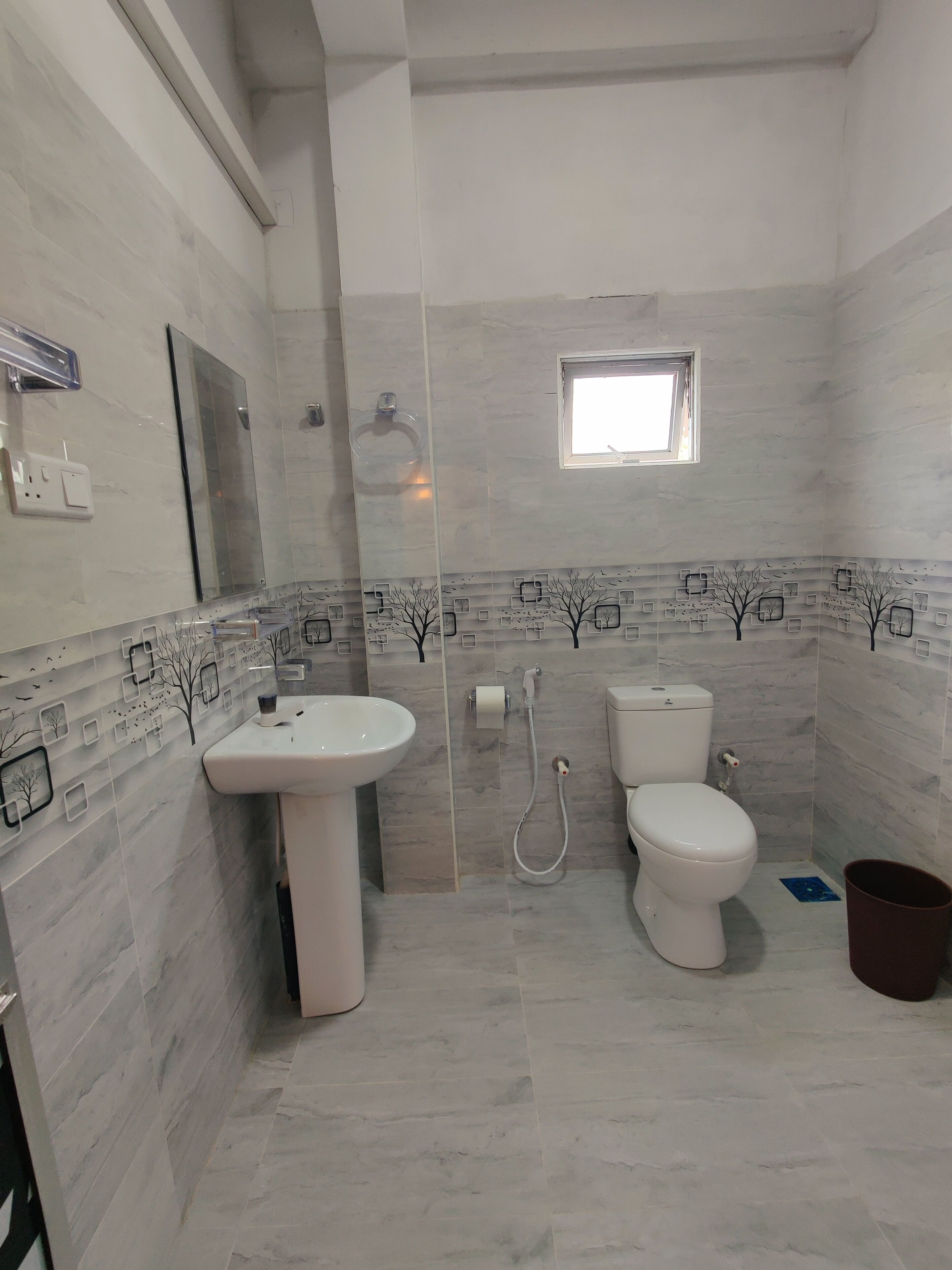 Un baño con lavamanos, inodoro y ducha. Hay un ventanal sobre el inodoro y un ventanuilla pequeña sobre la ducha. Las paredes tienen un diseño de árboles y cuadrados.