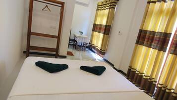 Habitación de hotel con una cama, un perchero de madera, una mesa con sillas, una planta en maceta y una cortina.