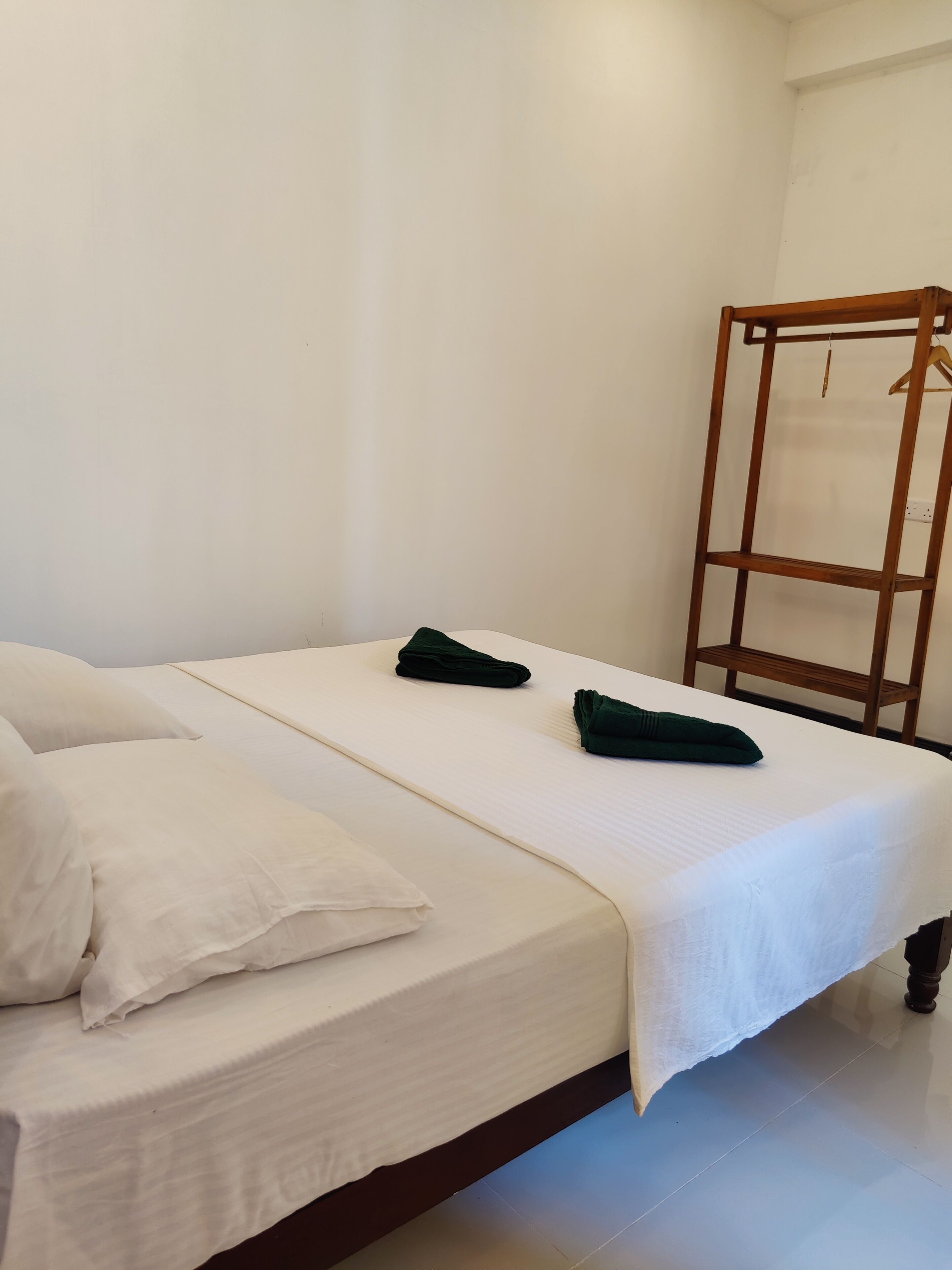 Een opgemaakt bed met witte lakens en twee groene handdoeken, een houten garderobe en een effen witte muur.