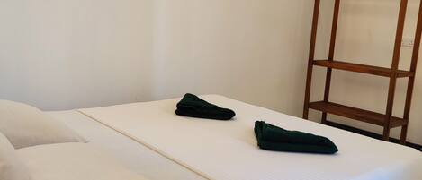 A neatly made bed with white linens and two green towels, a wooden wardrobe, and a plain white wall.