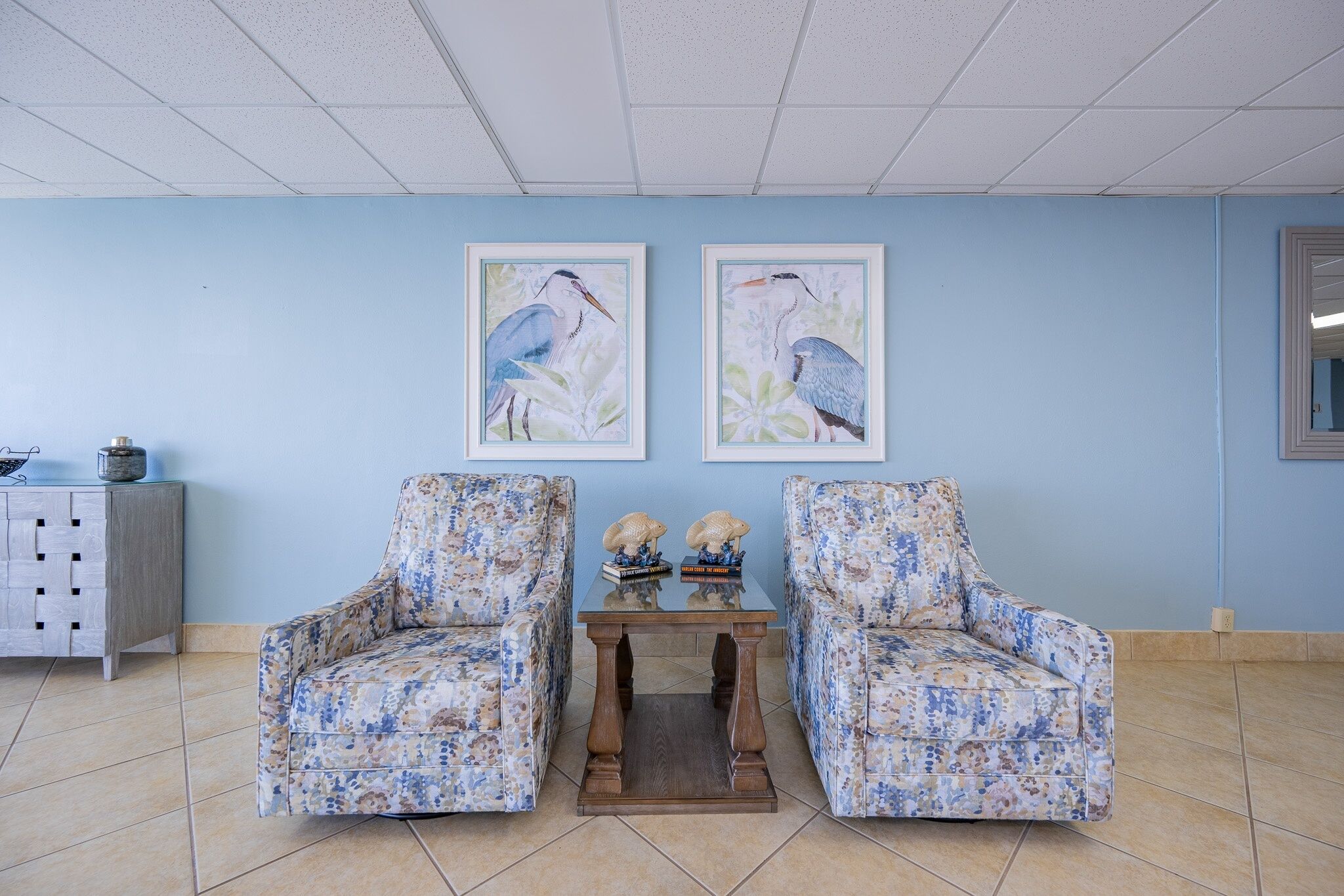 A room with two floral-patterned armchairs, a wooden table with decorative items, and framed bird paintings on the wall.