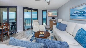 Una sala de estar con un sofá blanco, almohadas azules, una mesita de centro y un televisor. Cuenta con un balcón con vistas al océano.