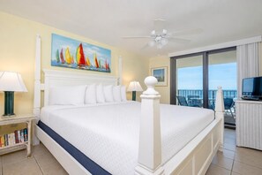 A hotel room with a large bed, a ceiling fan, a painting of sailboats, a television, and a balcony with chairs.