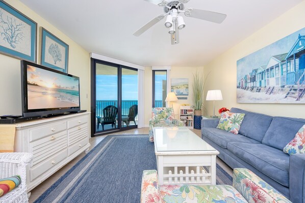 A living room with a television, sofa, coffee table, and chairs. There is a large window with a view of the ocean.