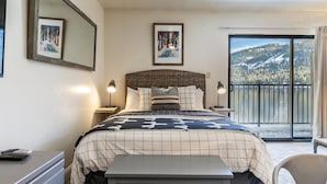 A hotel room with a bed, bedside lamps, a desk, and a chair. There are two framed pictures on the wall, and a balcony with a view of a snowy mountain. - 245 Bldg C 1 Bedroom Condo (Truckee)