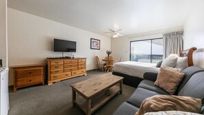 Habitación de hotel con cama, sofá, cómoda de madera, televisión, ventilador de techo y ventana con cortinas.