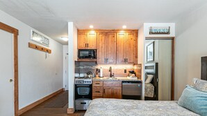 A compact kitchen with wooden cabinets, a microwave, stove, and dishwasher. A bed with patterned bedding is visible in the foreground. - 243 Bldg C 1 Bedroom Condo (Truckee)