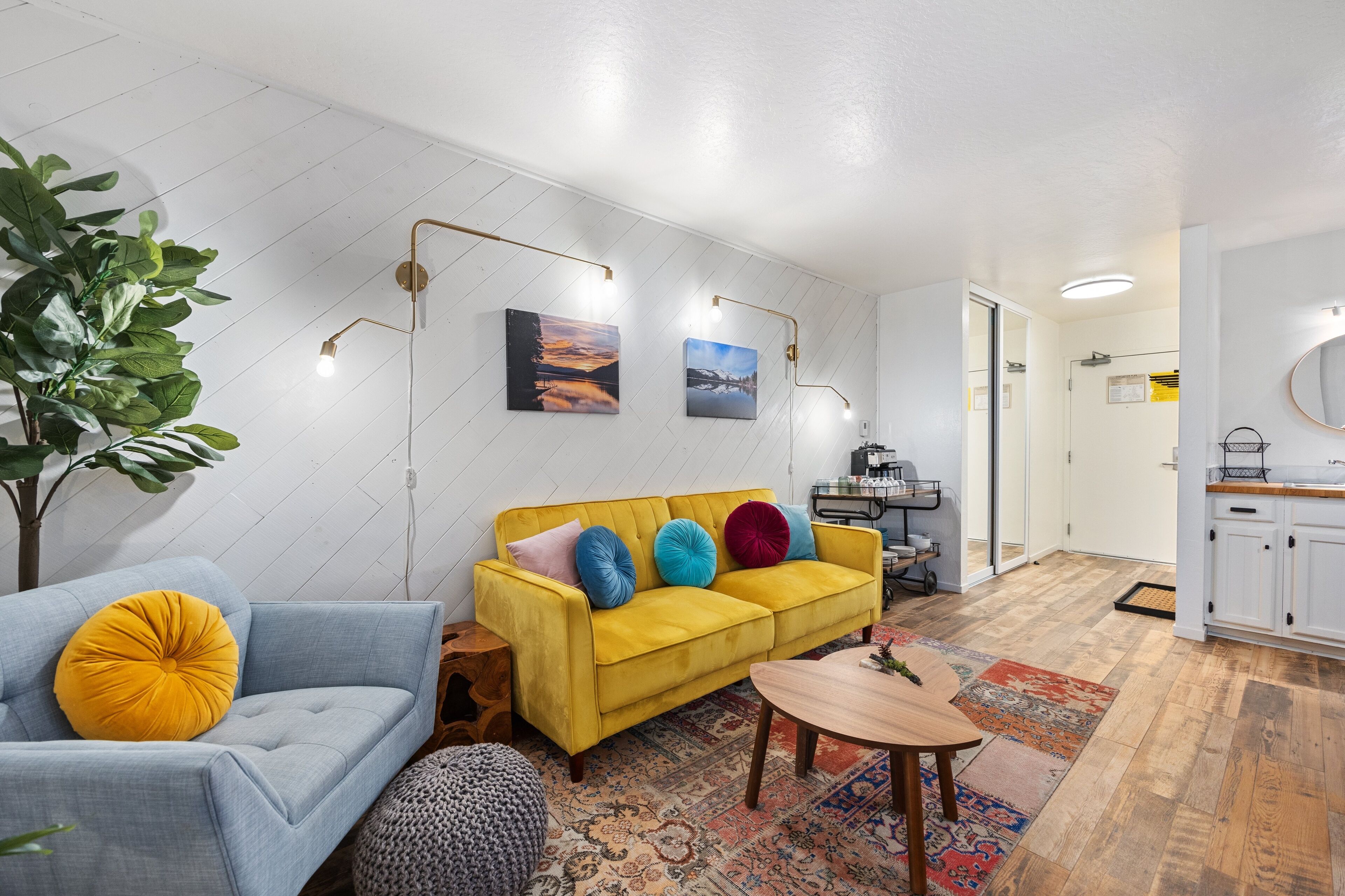 Una sala de estar moderna con un sofá amarillo, un sillón azul y una mesa de centro de madera. Hay dos lámparas fijadas a la pared y una planta grande.