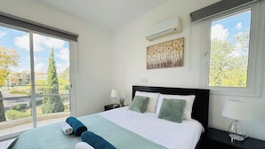 Un dormitorio con una cama grande, una ventana con vistas a árboles y edificios, un equipo de aire acondicionado y una mesita de noche con lámpara.