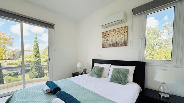 Un dormitorio con una cama grande, una ventana con vistas a árboles y edificios, un equipo de aire acondicionado y una mesita de noche con lámpara.