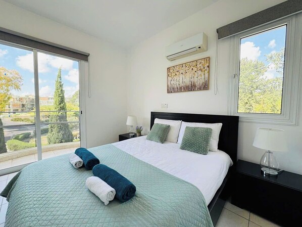 Un dormitorio con una cama grande, una ventana con vistas a árboles y edificios, un equipo de aire acondicionado y una mesita de noche con lámpara.