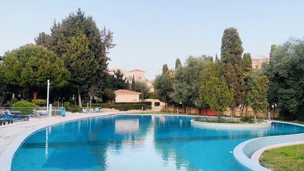 Una piscina de borde curvo, rodeada de árboles y un edificio al fondo.