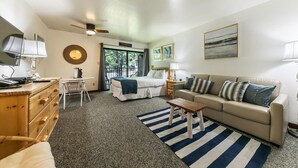 Habitación de hotel con cama, sofá, escritorio y una silla. Se ve un ventanal con vistas a árboles y un balcón.
