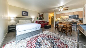 A compact hotel room with a bed, a red sofa, a dining table with chairs, a kitchen area with a microwave and stove, and a ceiling fan.