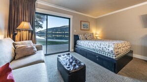 A hotel room with a sofa, a bed with patterned bedding, a small ottoman, a lamp, and a large window with a view of a lake and trees. - 146 Bldg C 1 Bedroom Condo (Truckee)