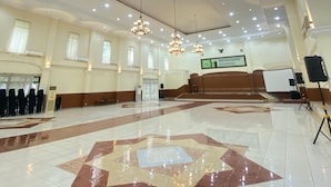 Ein geräumiger Saal mit einem glänzenden Fliesenboden, mehreren Deckenleuchten und einer Bühne mit Rednerpult.