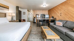 Quarto de hotel com cama, sofá, pequena mesa de jantar, cozinha compacta e painel de parede em madeira.