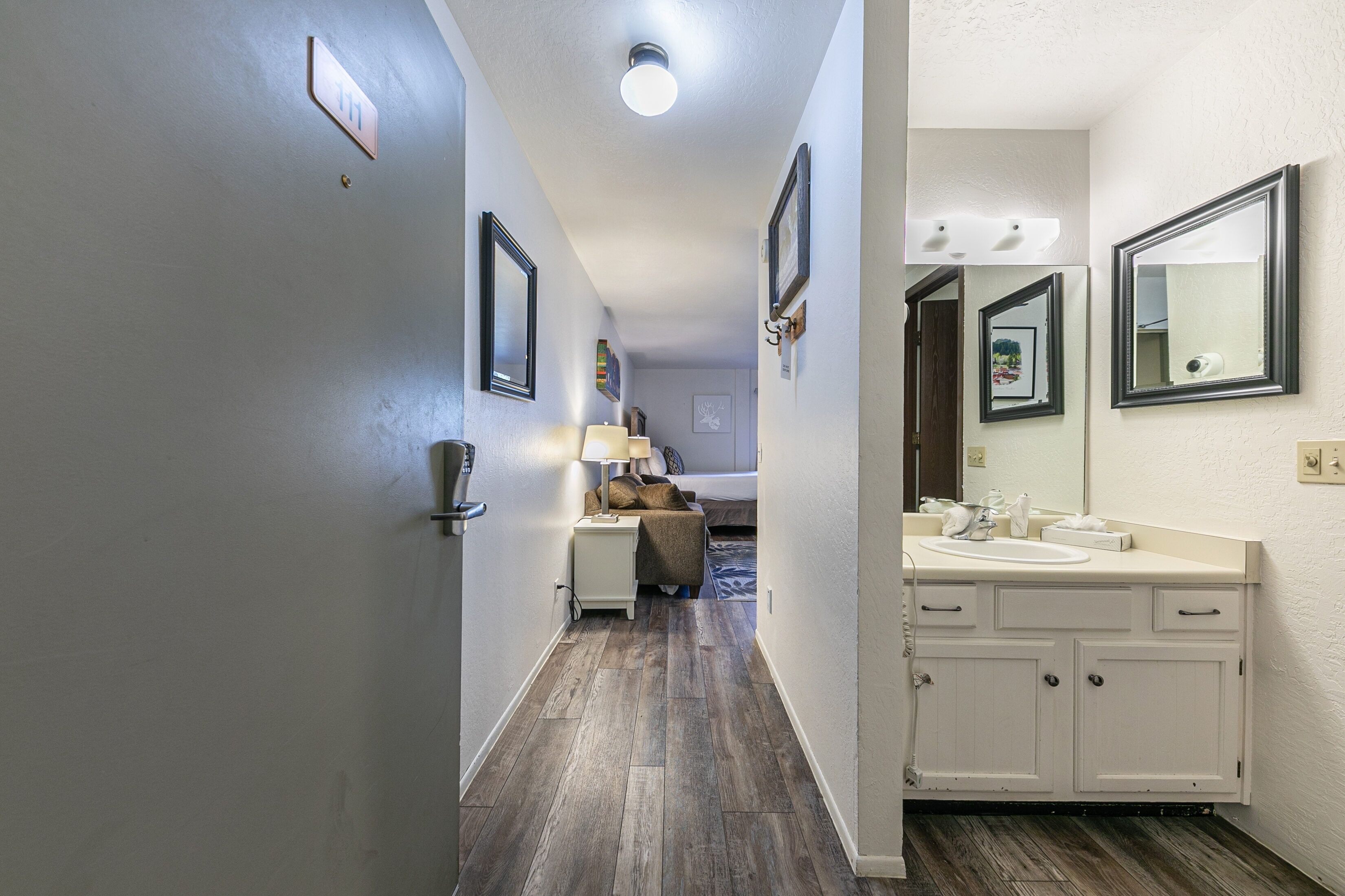 A hotel hallway with a wooden floor, a bed with a headboard, a nightstand with a lamp, and a bathroom with a sink and mirror.