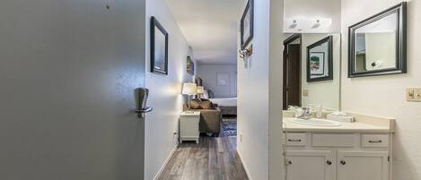 A hotel hallway with a wooden floor, a bed with a headboard, a nightstand with a lamp, and a bathroom with a sink and mirror.