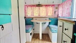 Cocina compacta con isla central, muebles blancos y una pequeña ventana con cortinas rosadas.