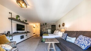 Un espace de vie compact comprenant un canapé, une petite table basse, un téléviseur fixé au mur, un coin cuisine et un lit recouvert d’une couverture verte.