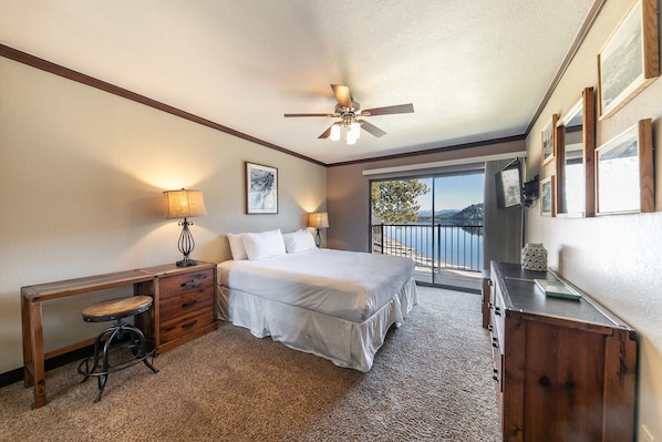 Habitación de hotel con una cama, dos lámparas de noche, un escritorio con una silla, un aparador y un balcón con vistas a un lago y montañas.