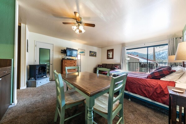 A cozy room with a wooden dining table, green chairs, a bed with red bedding, a TV, and a ceiling fan.