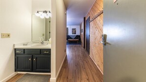 Un pasillo estrecho con suelo de madera, una puerta oscura a la derecha y un baño a la izquierda con lavabo y espejo.