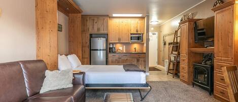 A compact living space with a bed, a brown leather sofa, a small wooden cabinet, and a kitchenette.