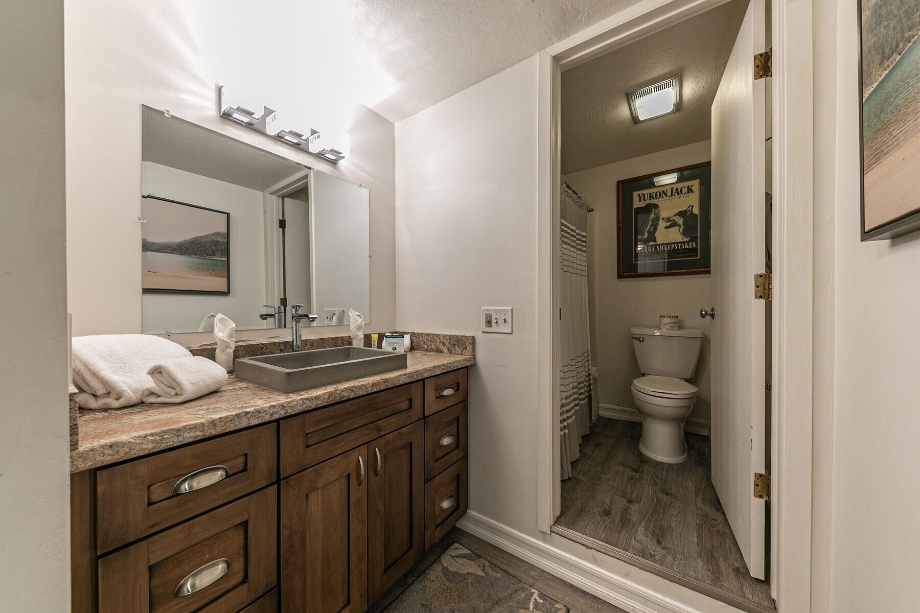A bathroom with a sink, mirror, and wooden cabinets. There is a poster on the wall and a door leading to another room.