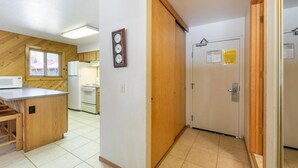 A compact kitchen with a microwave, stove, and wooden cabinets. A clock is mounted on the wall, and a door with a notice is visible. - 117 Bldg B 1 Bedroom Condo (Truckee)