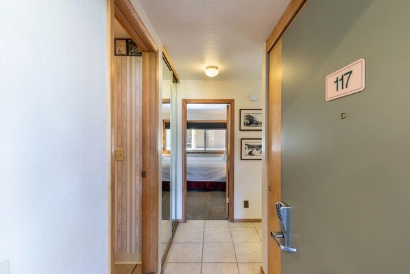 Un pasillo con una puerta de madera que tiene el número 117, que da a una habitación con cama y ventana.
