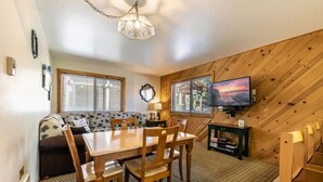 A cozy living room with a wooden wall, a sofa, a dining table with chairs, a television, and a fireplace. - 117 Bldg B 1 Bedroom Condo (Truckee)