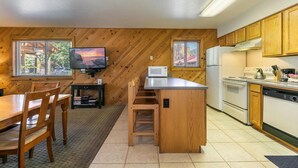 A compact kitchen with wooden cabinetry, a central island, a microwave, a stove, and a dishwasher. There is a dining area with a table and chairs, and a television mounted on the wall. - 117 Bldg B 1 Bedroom Condo (Truckee)